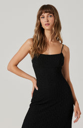 A woman is wearing a fitted, black, textured dress with a straight neckline and thin spaghetti straps.